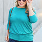 Curve model wearing a turquoise round neck batwing sleeve top, blue jeans, and sunglasses while walking outdoors.