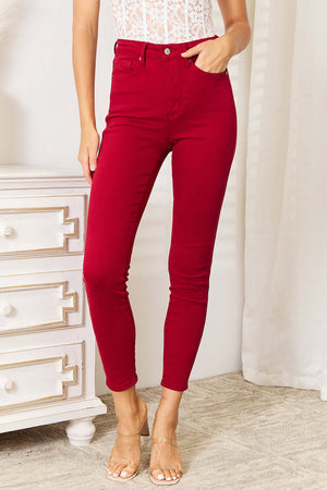 High Waist Tummy Control Skinny Jeans in Red by Judy Blue, offering a trendy silhouette and sleek look, shown on a model standing by a dresser