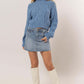 Model showcasing a blue cable-knit mock neck sweater with dropped shoulders, paired with a denim skirt and white knee-high boots.