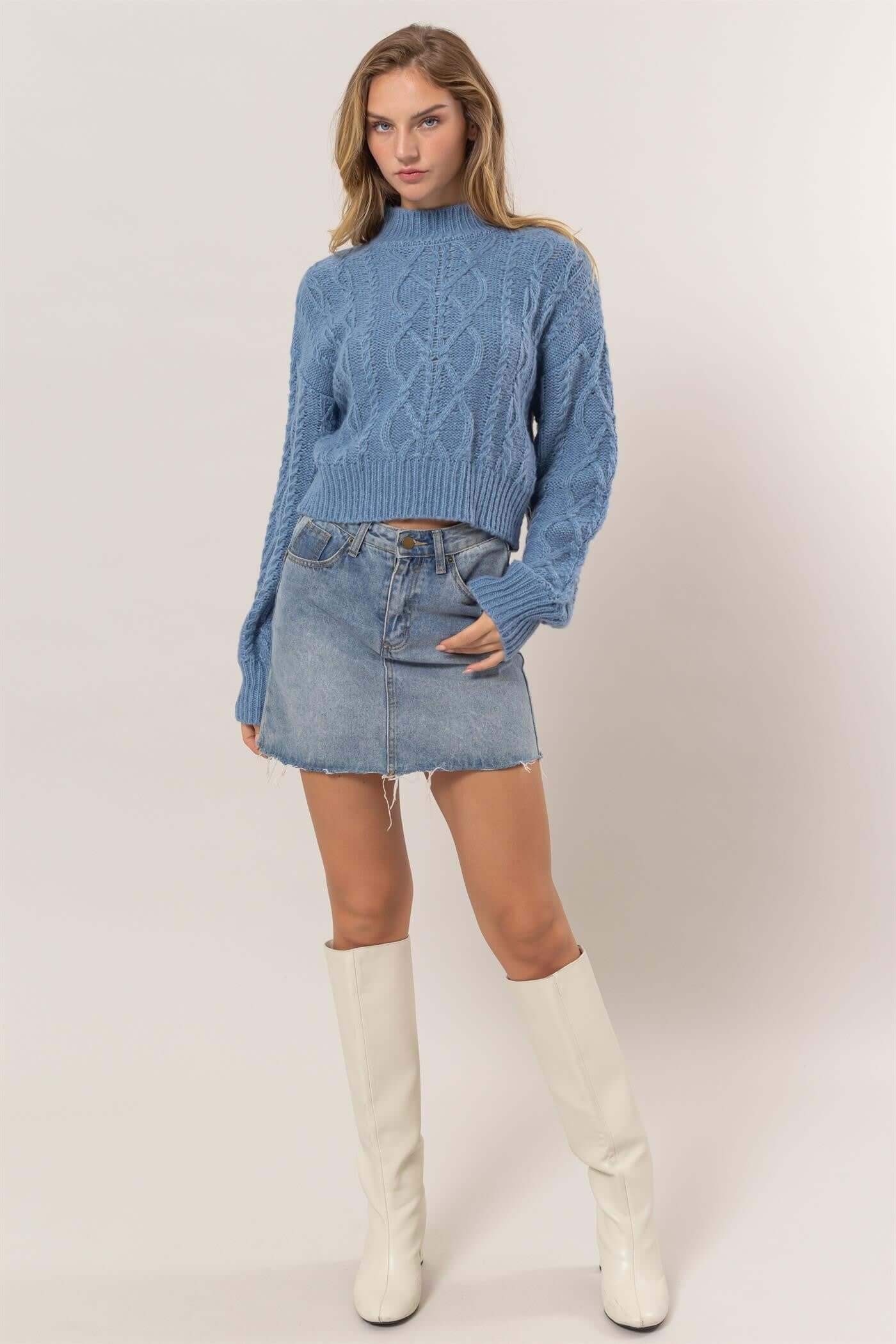 Model showcasing a blue cable-knit mock neck sweater with dropped shoulders, paired with a denim skirt and white knee-high boots.