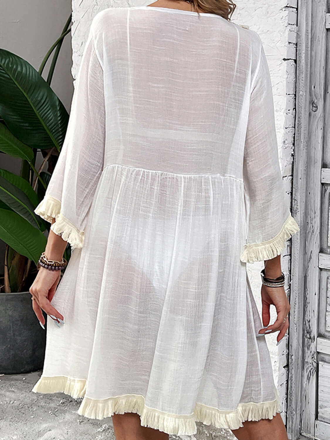 Woman wearing Fringe Tie Neck Three-Quarter Sleeve Cover Up, sheer white material with fringe details on sleeves and hem, back view.