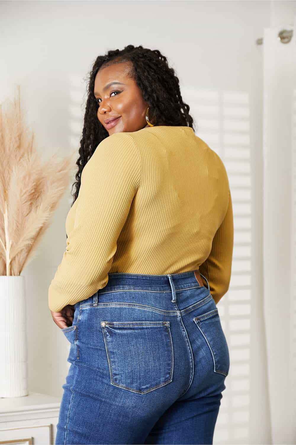 Woman wearing All About Details Full Size Ribbed Bodysuit in mustard yellow, paired with high-waisted blue jeans, showing back view.