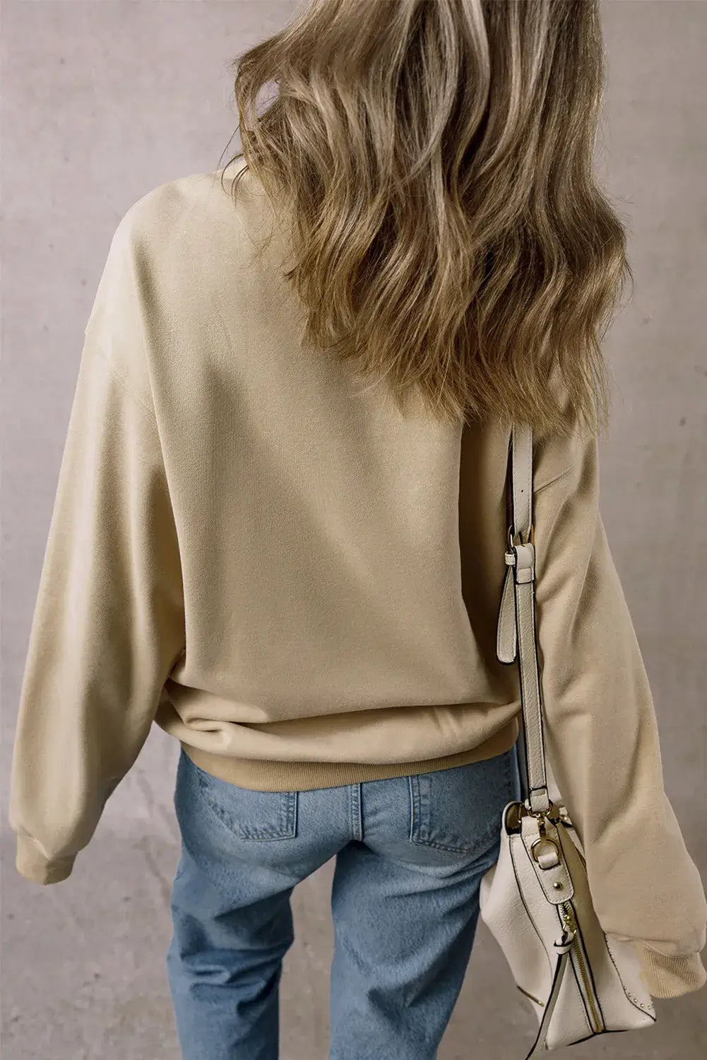 Woman wearing a Bella Road Thankful Round Neck Long Sleeve Sweatshirt in beige with blue jeans, showing back view holding a white handbag