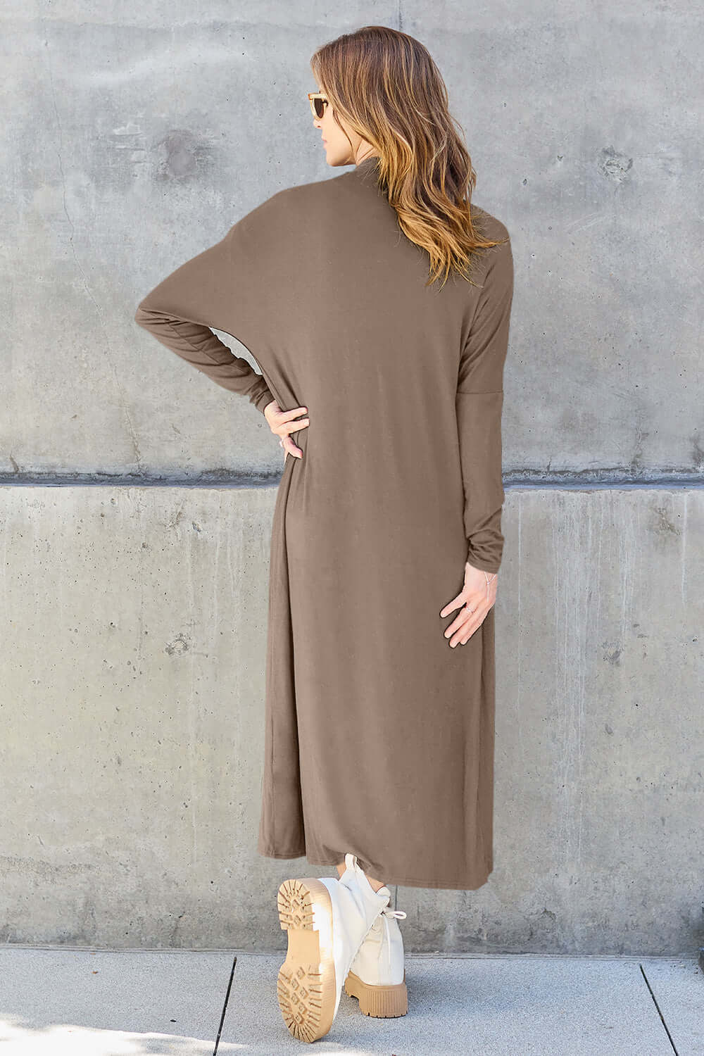 Woman wearing brown open front long sleeve cover-up with pockets, viewed from the back, standing against a concrete wall