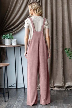 Woman wearing ribbed front pocket sleeveless jumpsuit in pink, viewed from the back, standing in a stylish, modern room with chic decor.