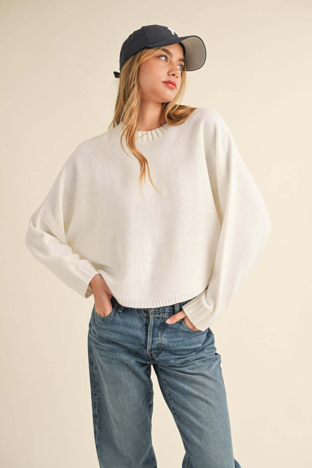 Woman wearing Bella Road Mable Round Neck Dolman Sleeve Cropped Sweater in white with blue jeans and a black baseball cap