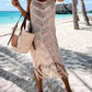 Model in a khaki fringe scoop neck cover-up by the beach, showcasing a stylish bohemian look with a tote bag.