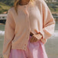 Woman in a soft pink Bella Road cardigan paired with striped shorts by the beach, perfect for cozy, stylish days.