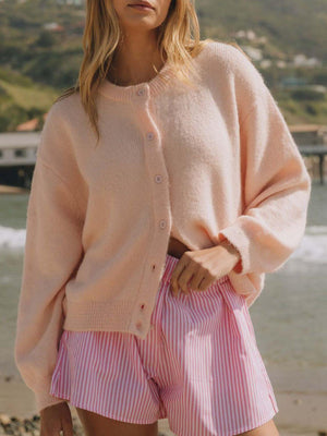 Woman in a soft pink Bella Road cardigan paired with striped shorts by the beach, perfect for cozy, stylish days.