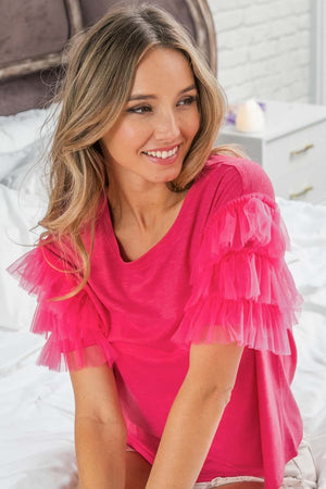 Woman wearing Ruffle Mesh Lace Layered Short Sleeve Top in bright pink, sitting on bed and smiling.