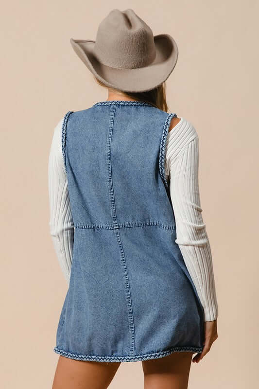 Back view of BiBi Braided Trim Open Front Denim Vest with Pockets, styled with a hat and layered over a long-sleeve top.