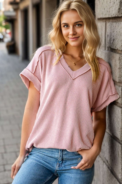 Woman wearing Exposed Seam Short Sleeve Ribbed Top in light pink, paired with blue jeans, standing outdoors by a brick wall.