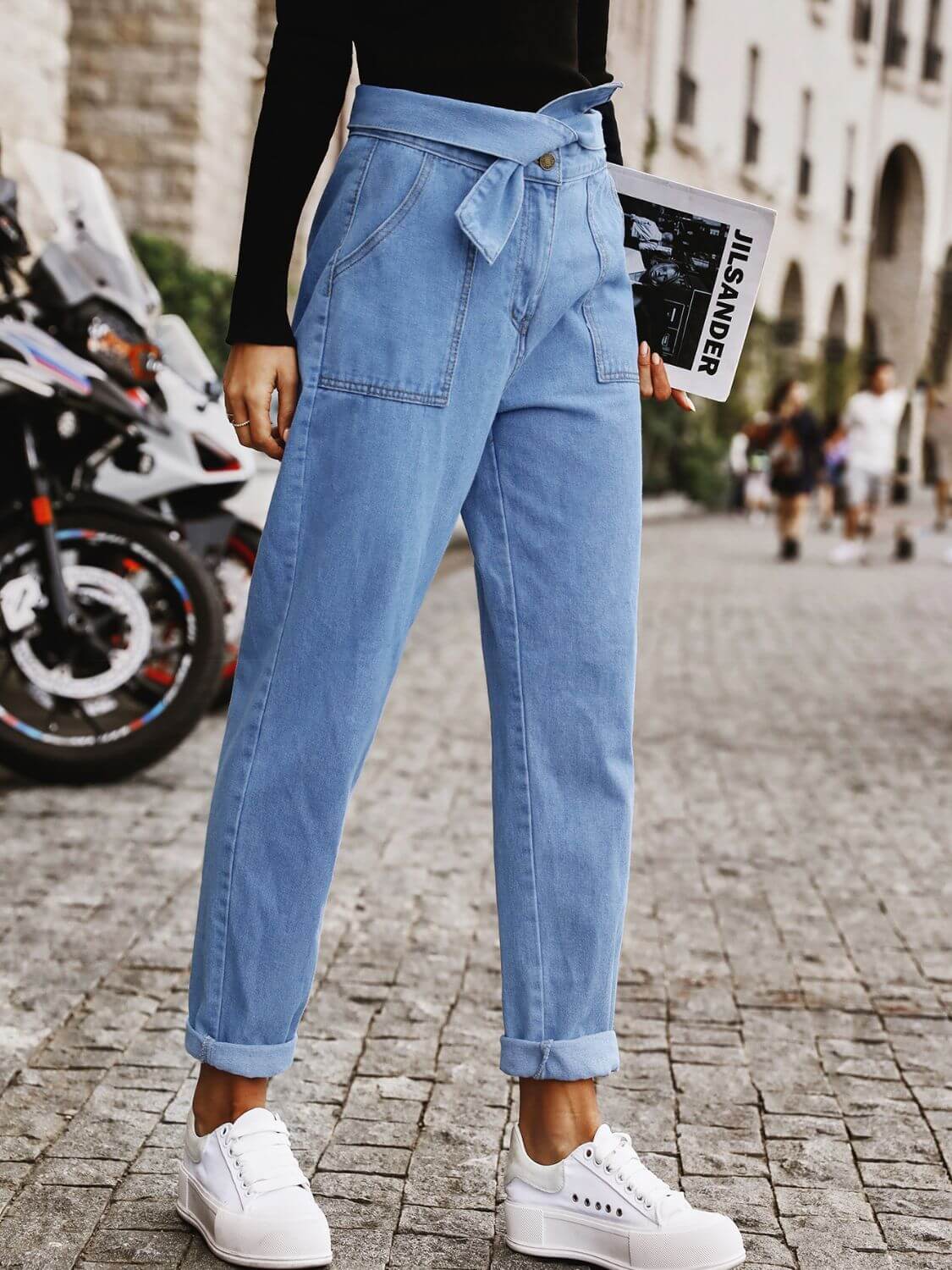 Woman wearing Bella Road Tied Straight Jeans with Pockets and white sneakers, holding a magazine while standing on a cobblestone street.