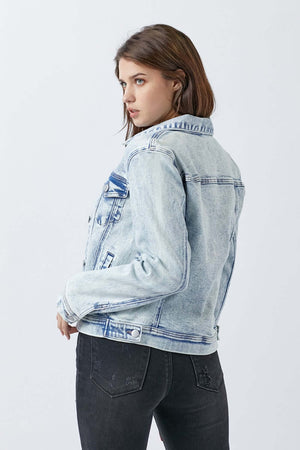 Woman wearing a button-up washed denim jacket by Risen Jeans, showing the back with classic buttoned chest pockets, paired with black jeans