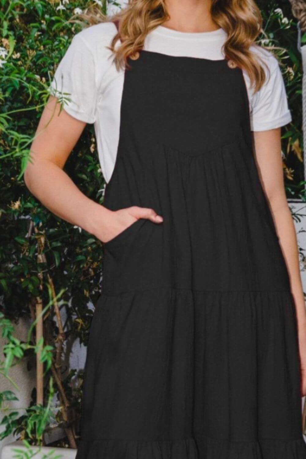 Woman wearing a sleeveless tiered midi dress in black, standing outdoors with greenery in the background.