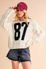 Stylish model wearing Aemi+Co Contrast Number Cable Knit Drop Shoulder Sweater with sporty 87 design and casual shorts.