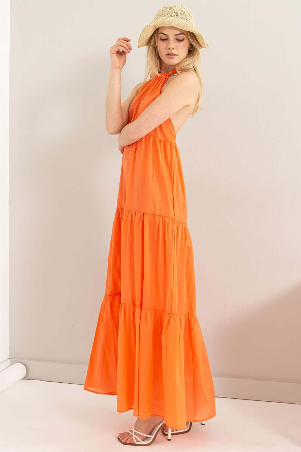 Model wearing a bright orange halter neck maxi dress with a tiered silhouette and a straw hat, perfect for summer outings.