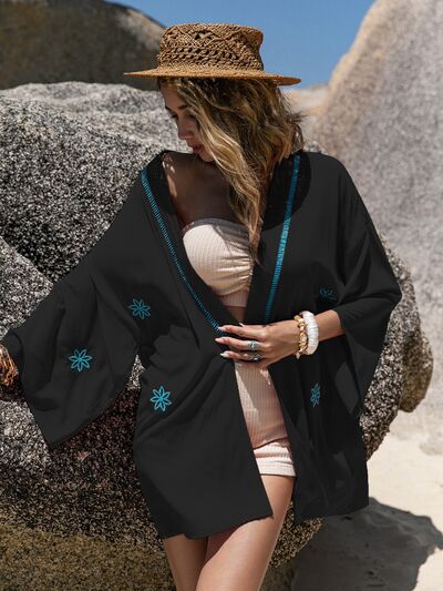 Woman in black Bella Road Swim cover-up by the beach, featuring vivid embroidery and stylish open front for summer vibes.