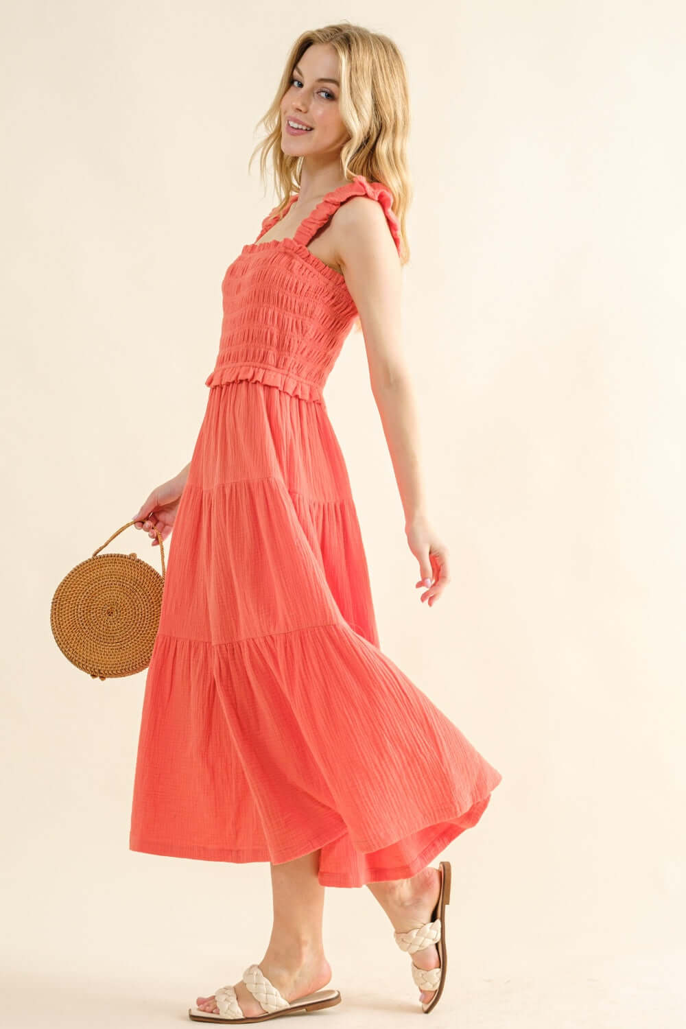 Woman wearing a coral smocked ruffled tiered dress holding a round straw bag.