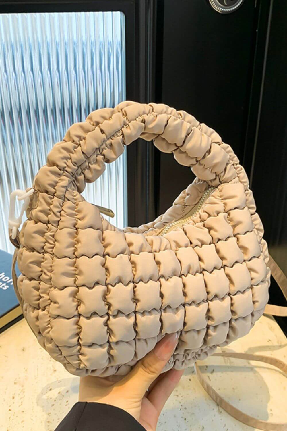 Beige quilted puffy crossbody bag with removable strap, held by a hand, showcasing stylish design and texture for versatile fashion use.