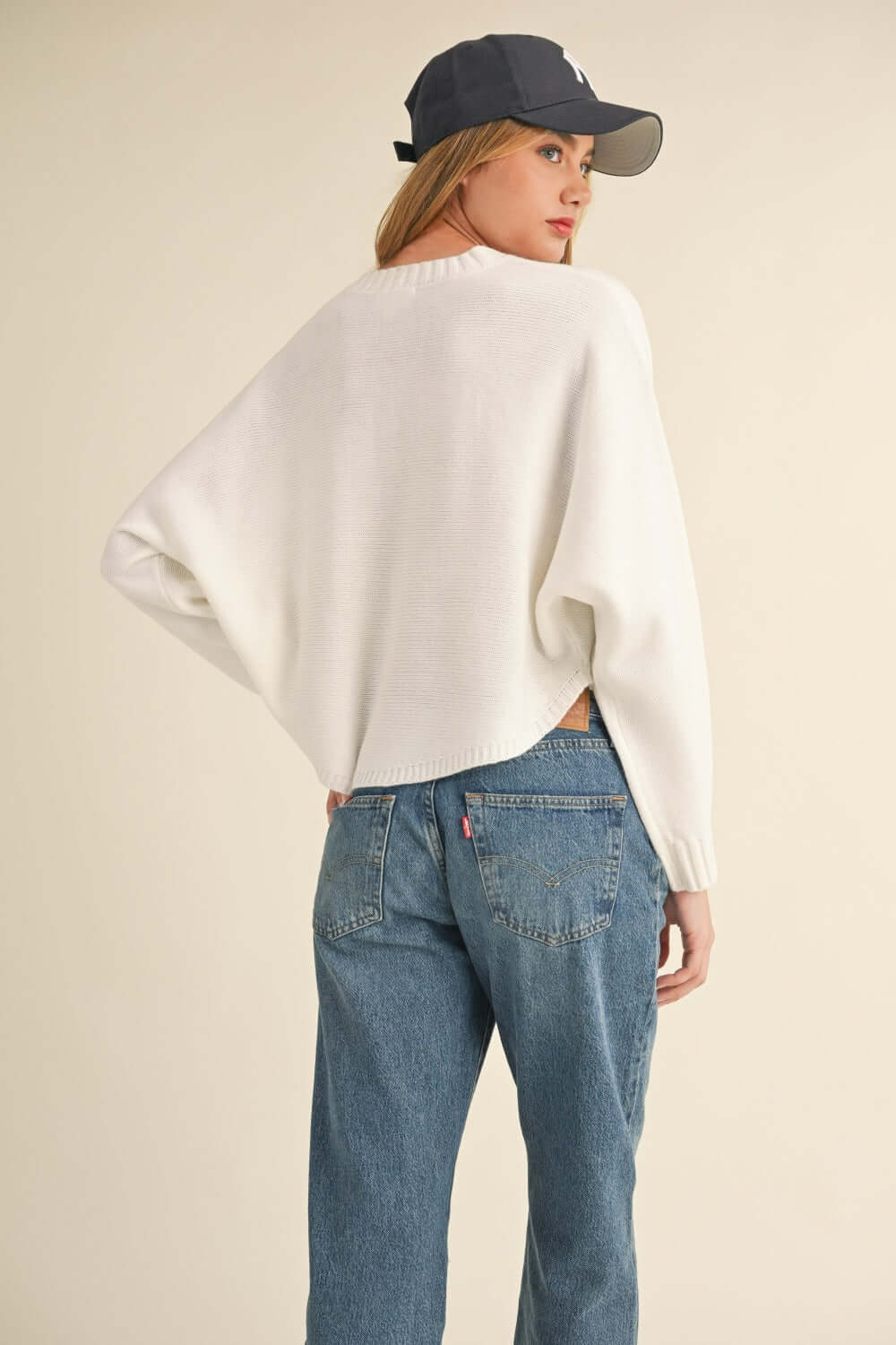 Woman wearing Bella Road Mable Round Neck Dolman Sleeve Cropped Sweater paired with jeans and a baseball cap, showing back view.