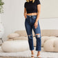 Woman wearing RFM Crop Dylan Full Size Tummy Control Distressed High Waist Raw Hem Jeans standing in a stylish living room.