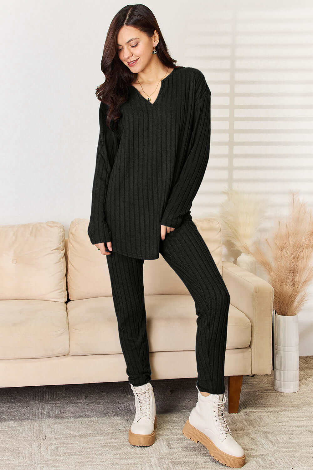 Woman wearing black notched long sleeve top and pants set, slightly stretchy ribbed two-piece outfit, standing next to a beige sofa.