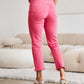 Woman in pink RFM jeans with tummy control and raw hem design, shown from behind in a stylish living room.