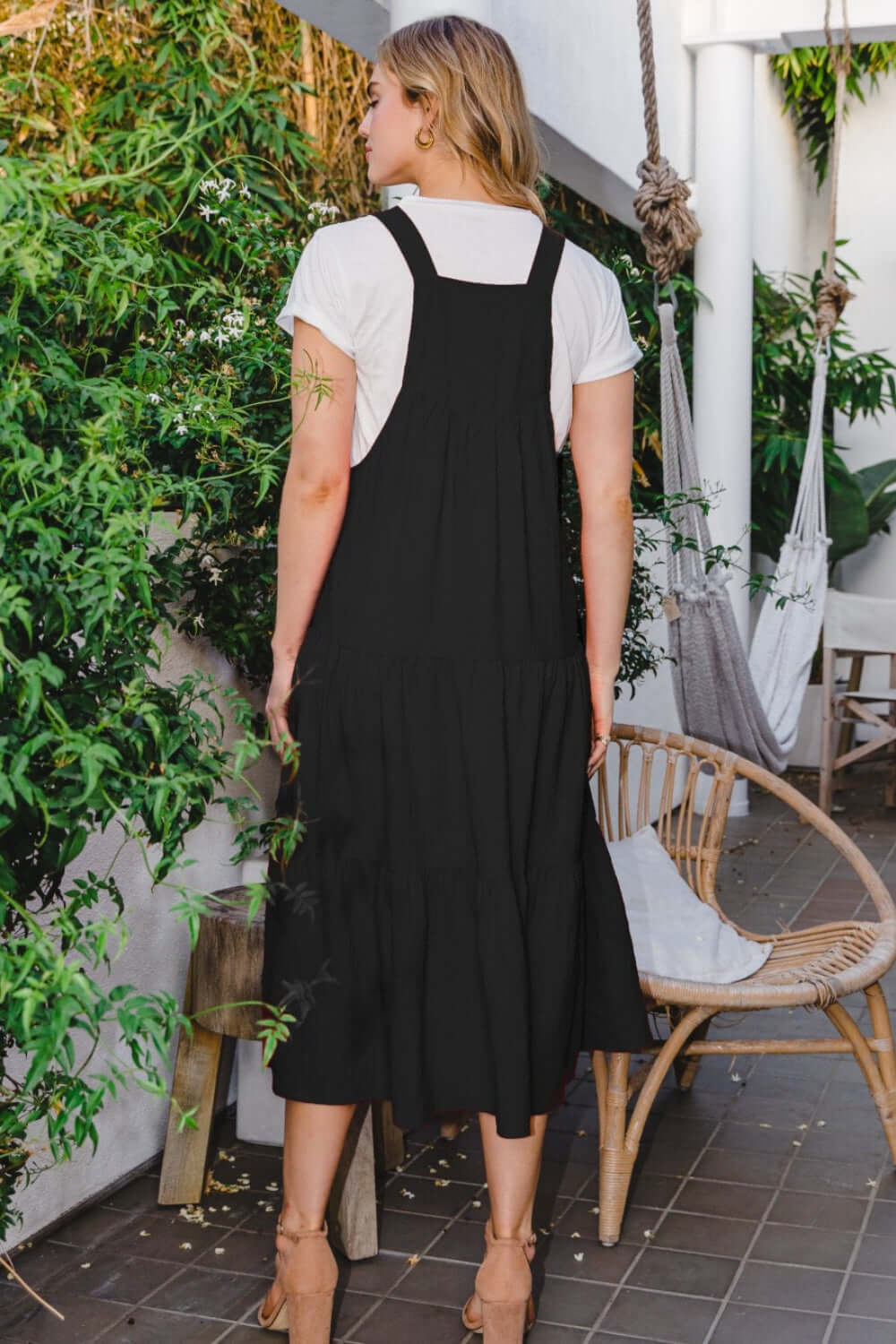 Woman wearing Sleeveless Tiered Midi Dress in black standing on a terrace surrounded by greenery.