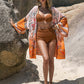 Model in Bella Road Swim printed cover-up over bikini, standing on beach with sun hat, surrounded by rocks.