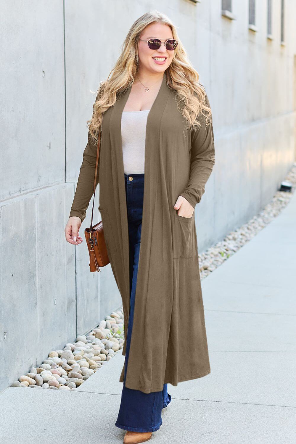 Woman wearing an open front long sleeve cover up with pockets in a taupe color, paired with wide-leg jeans and a white top, walking outdoors.