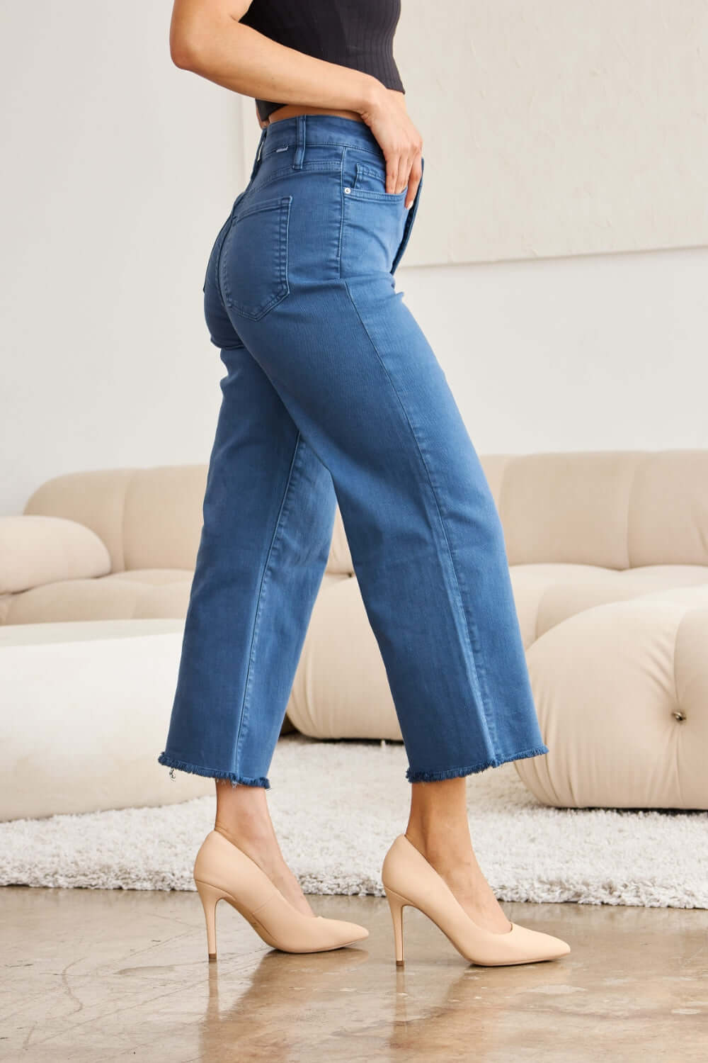 Woman wearing Crop Chloe Tummy Control High Waist Raw Hem RFM Jeans with heels in a stylish indoor setting