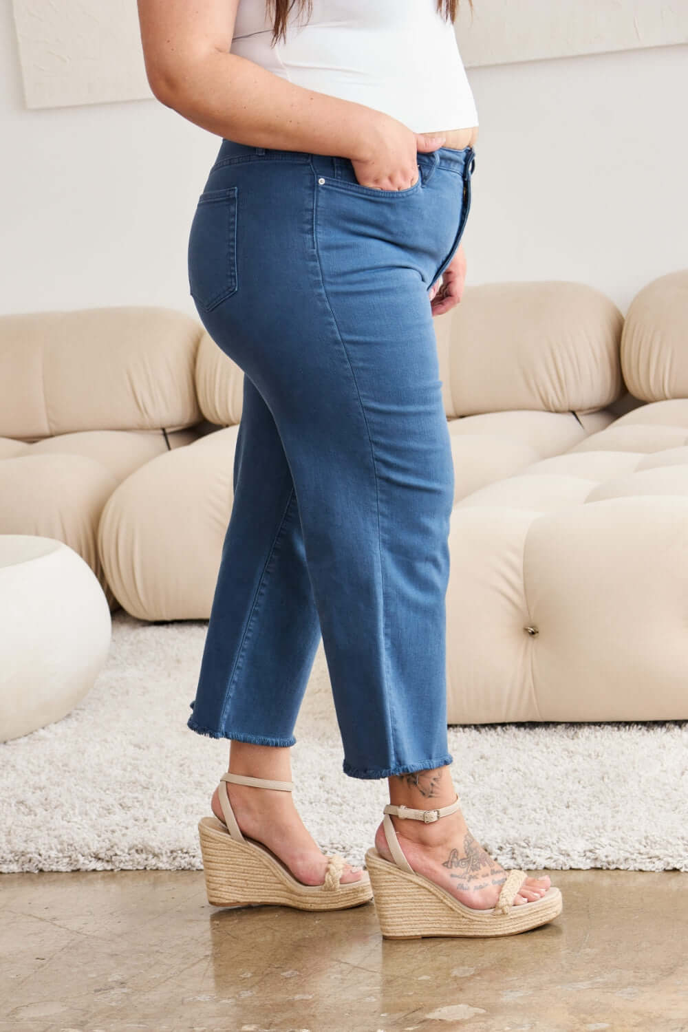 Woman wearing RFM Jeans Tummy Control High Waist Raw Hem, posing in living room with wedge sandals