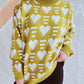 Woman wearing Bella Road Heart Contrast Long Sleeve Dropped Shoulder Sweater in yellow with white heart pattern, standing against a wall.