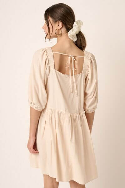 Back view of a chic beige mini dress with pintuck detail and tie-back design, showcasing playful puff sleeves.