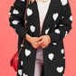 Woman wearing Bella Road Heart Open Front Long Sleeve Cardigan with white hearts, holding a red purse against a pink background.