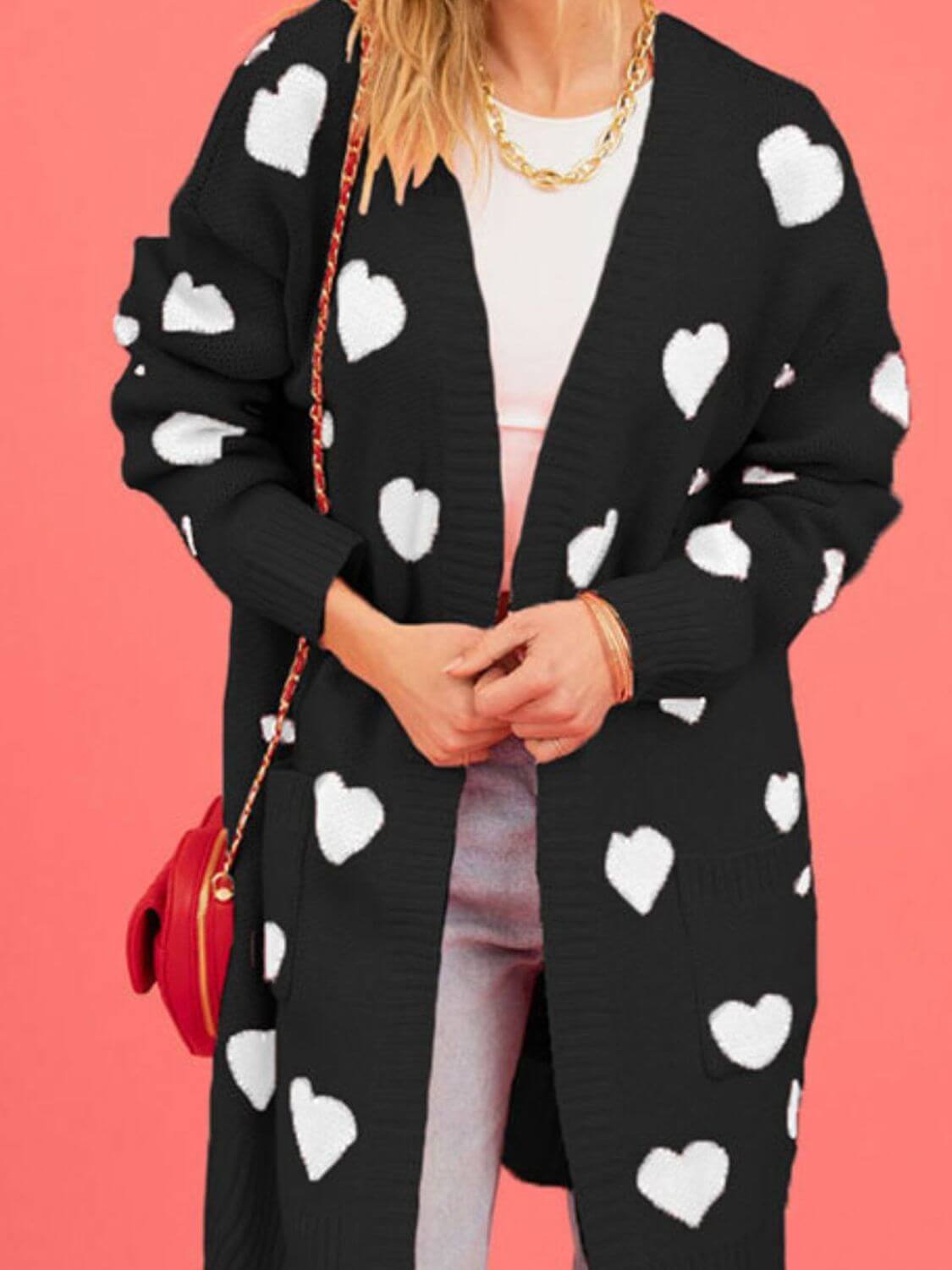 Woman wearing Bella Road Heart Open Front Long Sleeve Cardigan with white hearts, holding a red purse against a pink background.