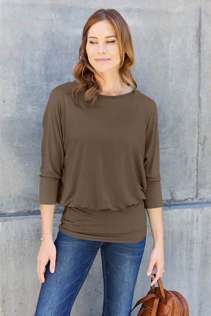 Woman wearing brown round neck batwing sleeve top with jeans, holding a brown handbag, standing against a concrete wall.