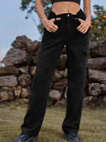 Woman wearing black high waist cargo straight jeans outdoors with hands in pockets against stone wall background.