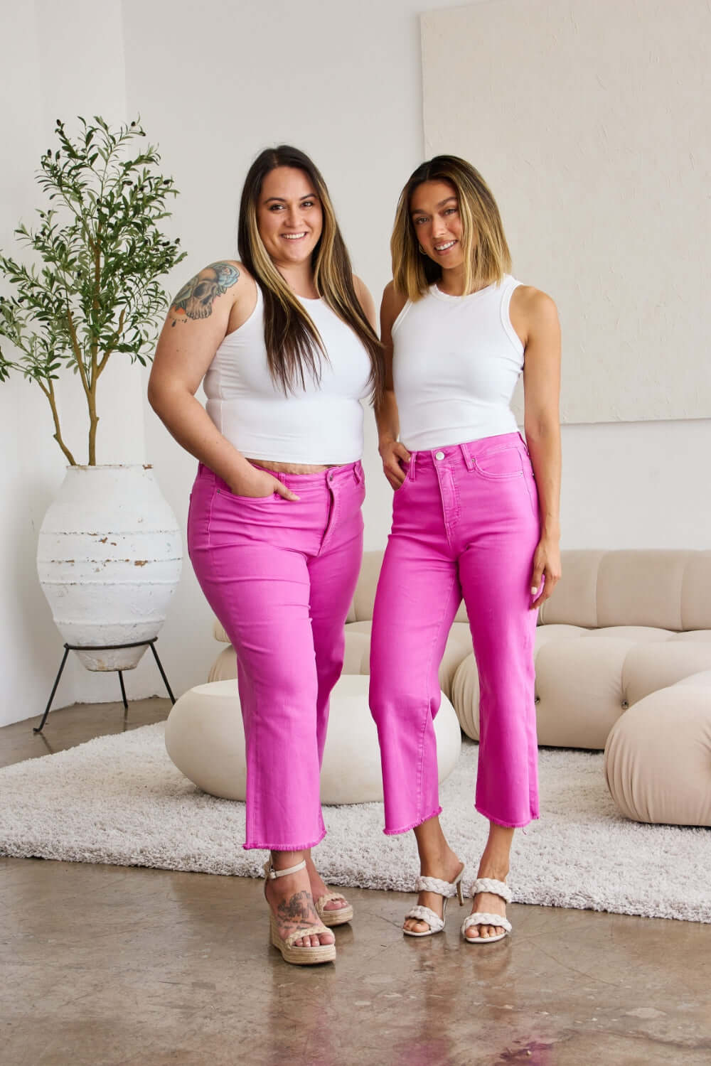 Two women wearing bright pink RFM Jeans with white tops, standing together in a stylish living room setting.