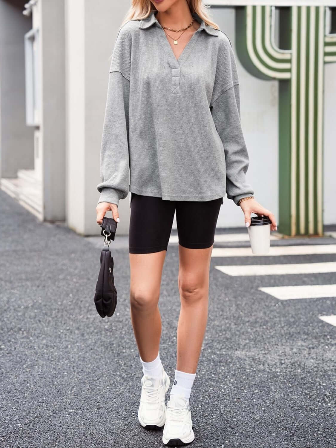 Woman wearing a grey waffle-knit Johnny collar long sleeve sweatshirt paired with black shorts and white sneakers, holding a coffee cup.