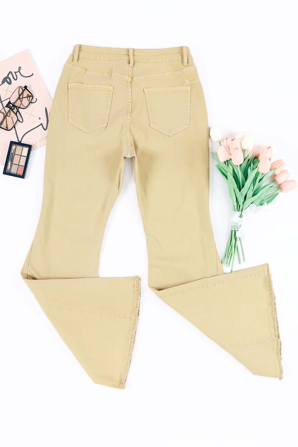 Back view of Bella Road Pocketed Raw Hem Flare Jeans in beige with flowers and makeup on the side.