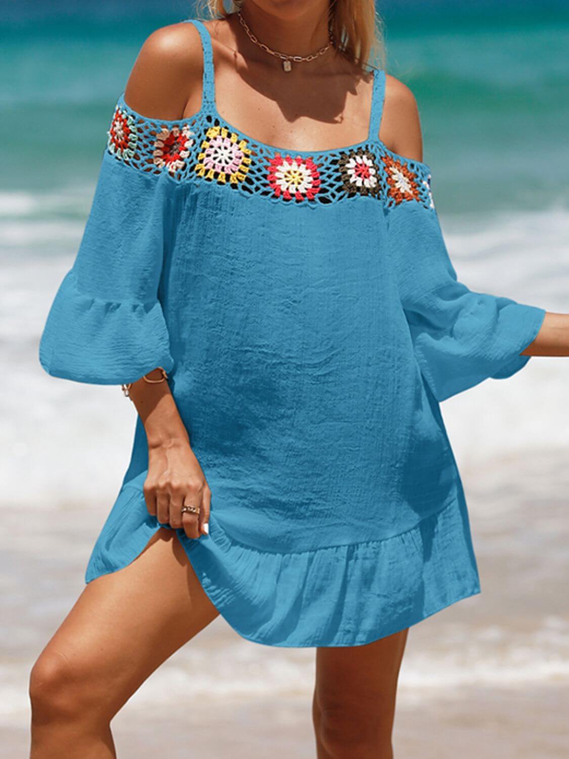 Woman wearing blue crochet cold shoulder three-quarter sleeve cover up with ruffled hem and colorful crochet details at the beach