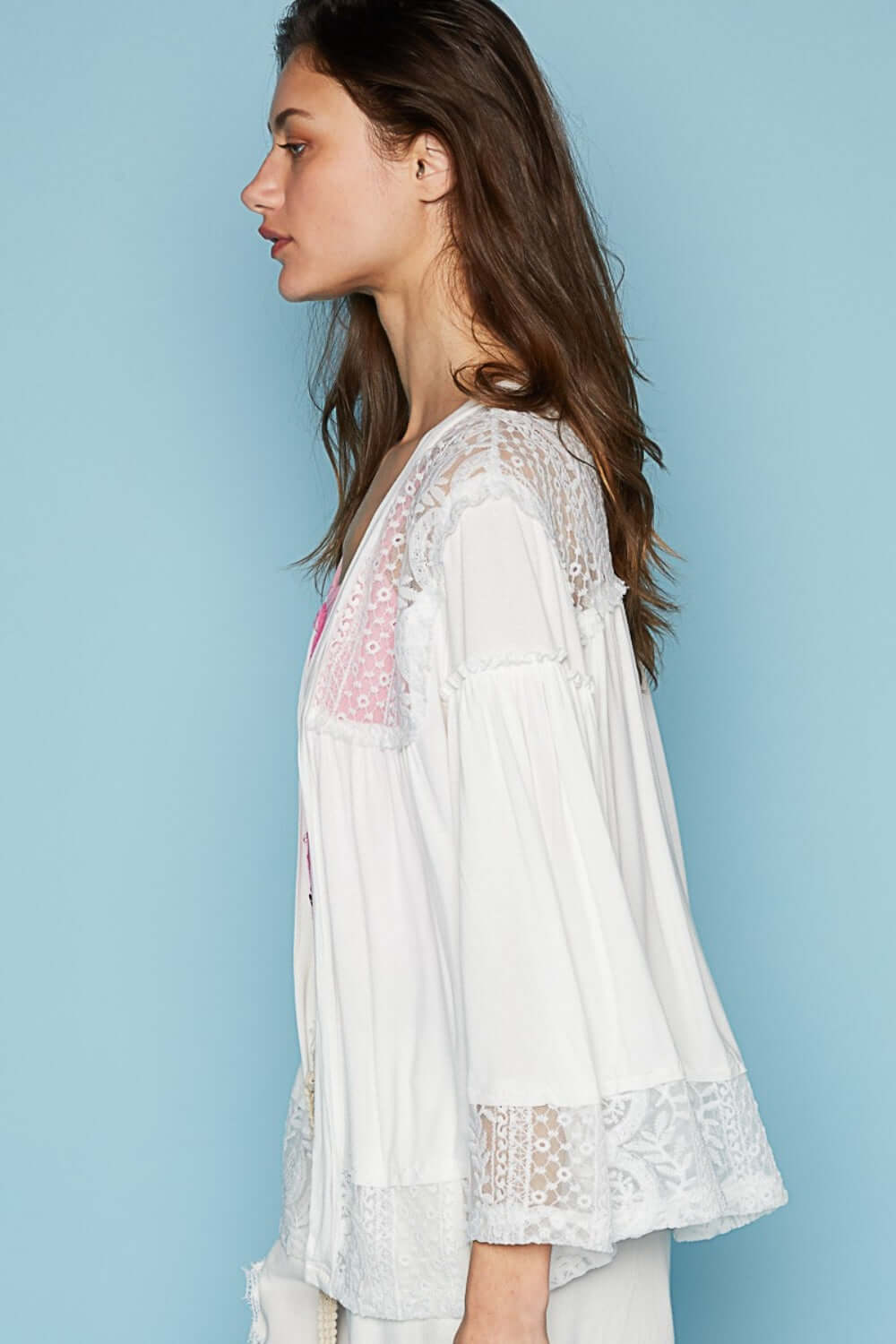 Woman wearing open front lace detail cardigan with 3/4 bell sleeves and frayed edges in a mix of jersey and lace, side view against blue background.