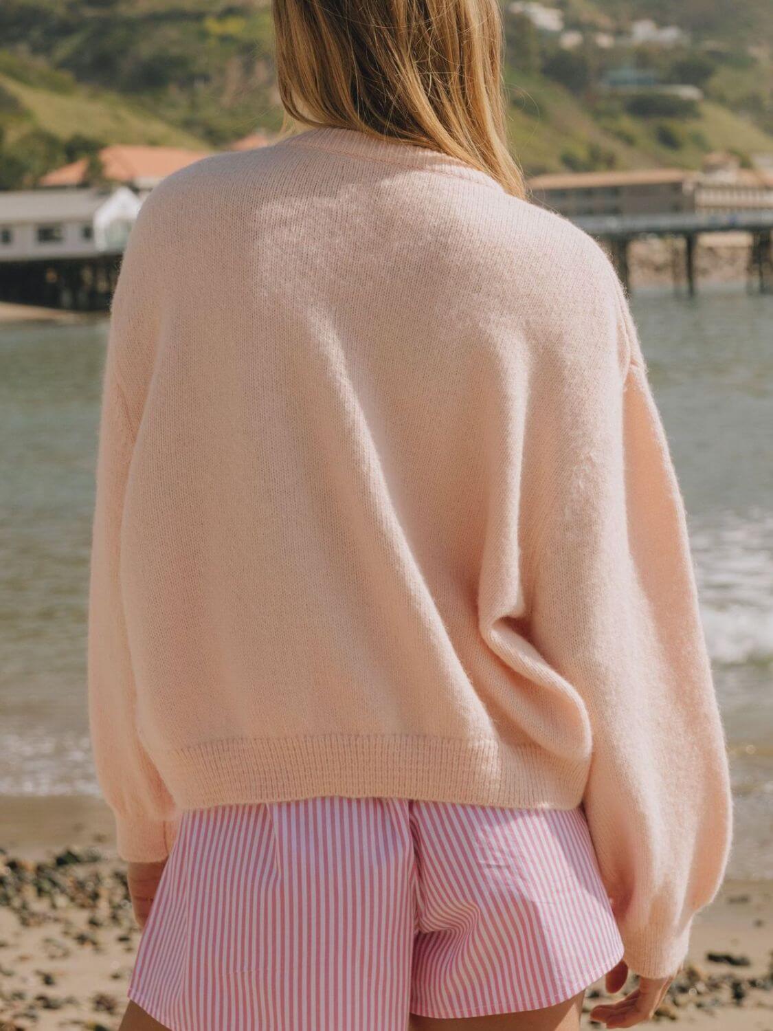 Woman in a cozy pink cardigan, enjoying a sunny day by the beach, wearing striped shorts.