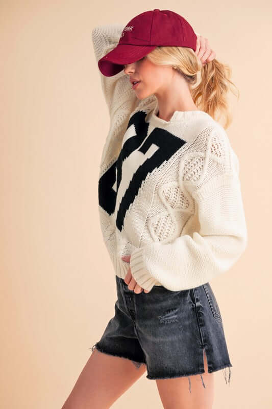 Model wearing Aemi+Co Contrast Number Cable Knit Sweater with black numbers, paired with denim shorts and a maroon cap.