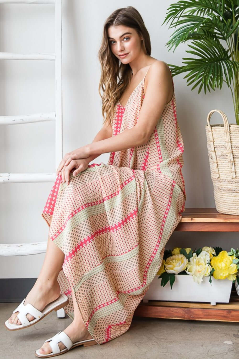 Woman wearing a vibrant print maxi cami dress with pockets, sitting on a bench next to a basket and flowers.