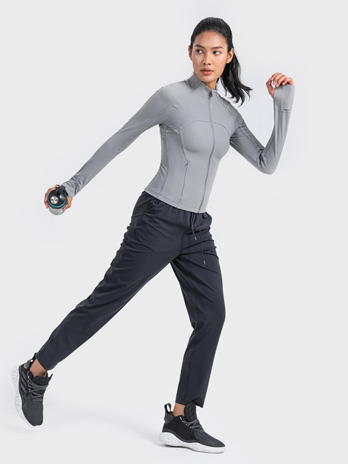 Woman in a gray zip-up long sleeve sports jacket, stylishly exercising with a dumbbell in hand. Perfect for any active lifestyle!