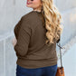 Woman wearing a brown round neck batwing sleeve top, blue jeans, and sunglasses, holding a brown handbag, smiling outdoors.