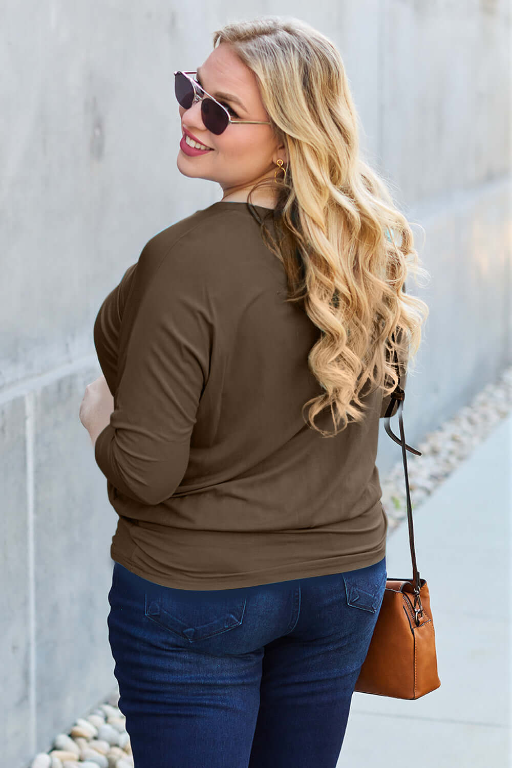 Woman wearing a brown round neck batwing sleeve top, blue jeans, and sunglasses, holding a brown handbag, smiling outdoors.